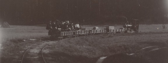 Svartvitt foto av Lillbässen, ett litet ånglok med fyra vagnar – lastade föremål och välklädda passagerare i den sista vagnen. Rök stiger från loket på en solig sommardag. Bild från Fiskars museums samlingar.