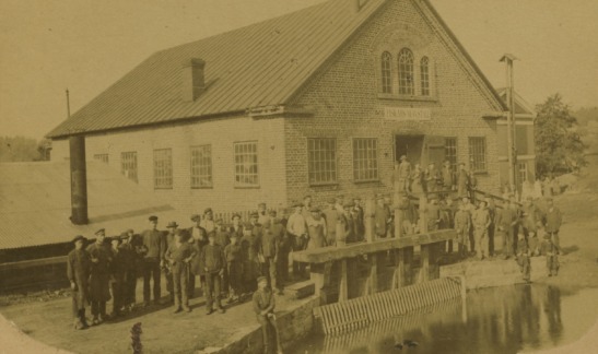 Svartvit fotografi av 53 arbetare vid Fiskars Mekaniska Werkstad, år okänt. Arbetarna poserar framför verkstaden i sina arbetskläder, vilket speglar industrisamhällets vardag. Fiskars Mekaniska Werkstad var den första platsen i Finland som producerade ångmaskiner, och bilden illustrerar dess betydelse för den finländska industrins utveckling. Från Fiskars Museums samlingar.
